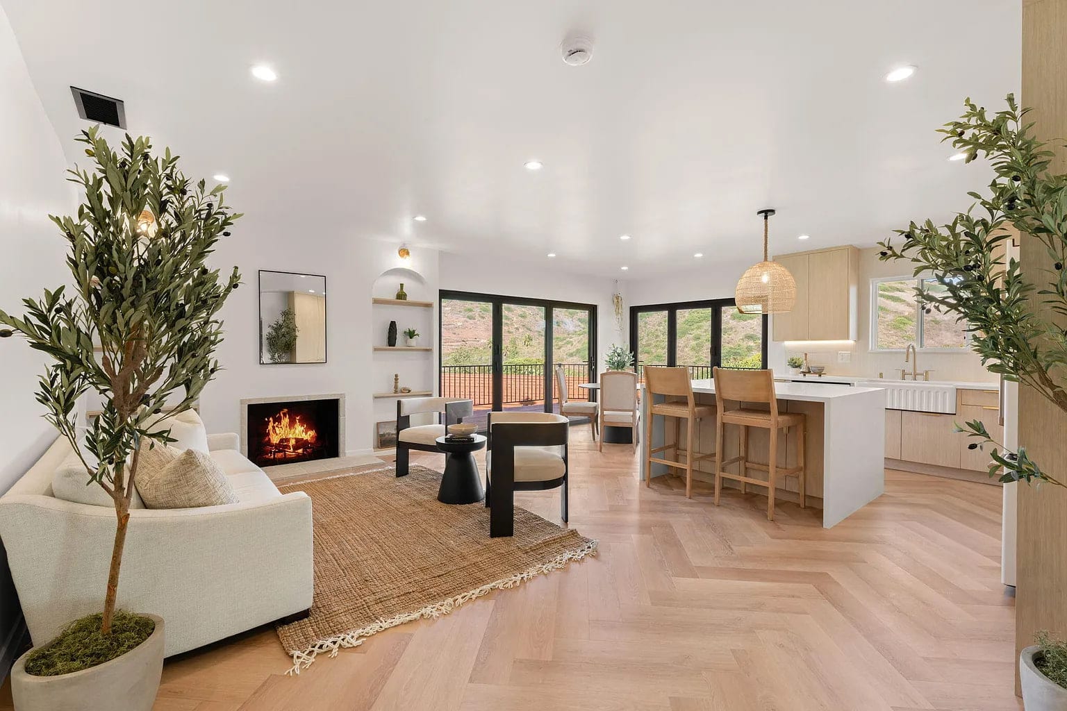 Open living and kitchen space with a lit fireplace, jute rug, and glass doors to a hillside view, DHRH Campanil renovation