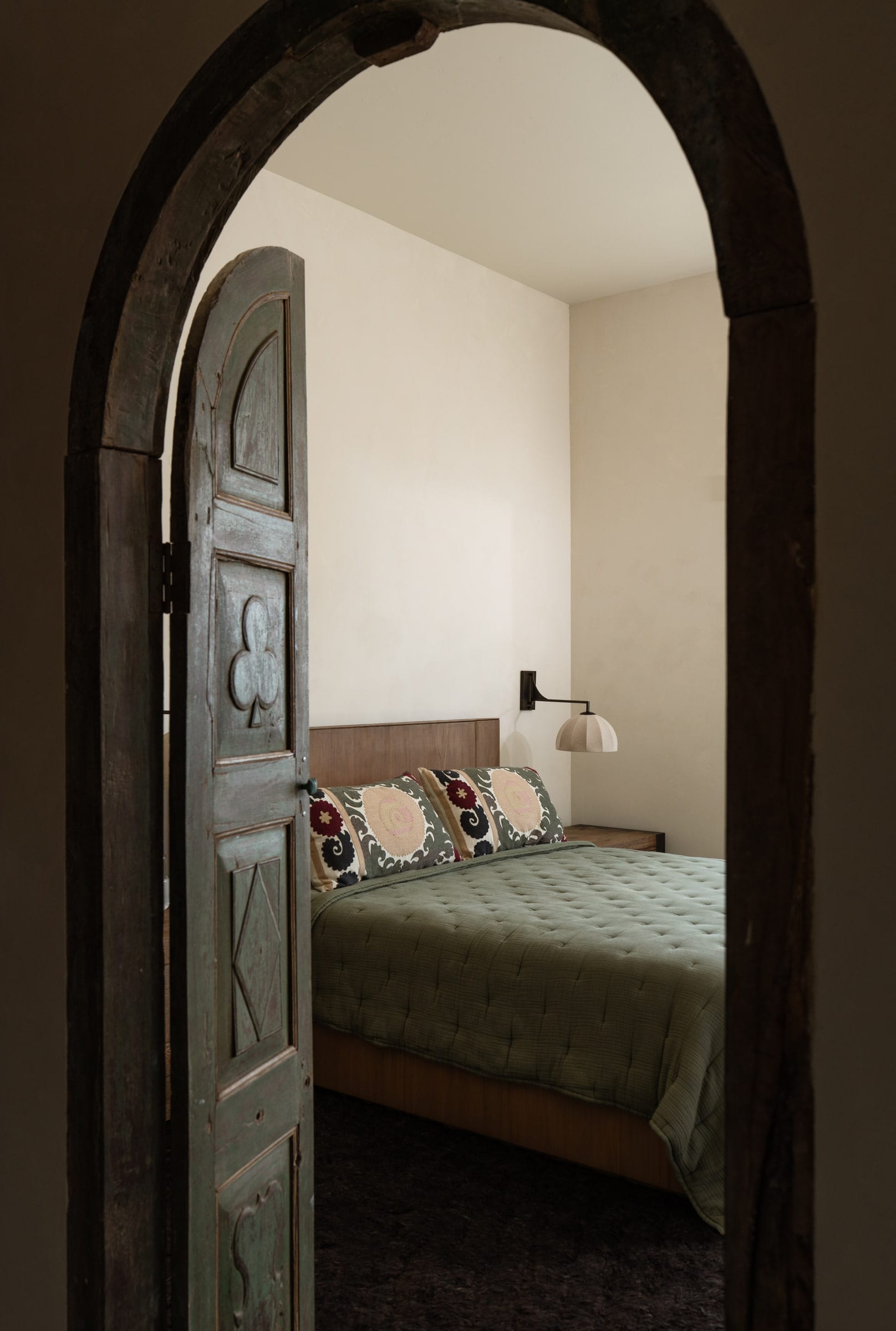 Bedroom viewed through an antique carved wood arched door, with a walnut headboard and sage quilted bedding, DHRH Mission Canyon renovation