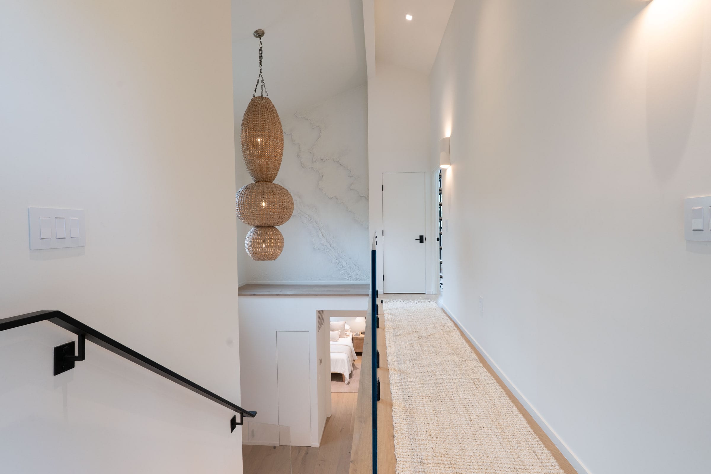 Upstairs landing with a tiered woven rattan pendant overlooking a bedroom below and a black metal stair rail, DHRH Padaro Lane renovation