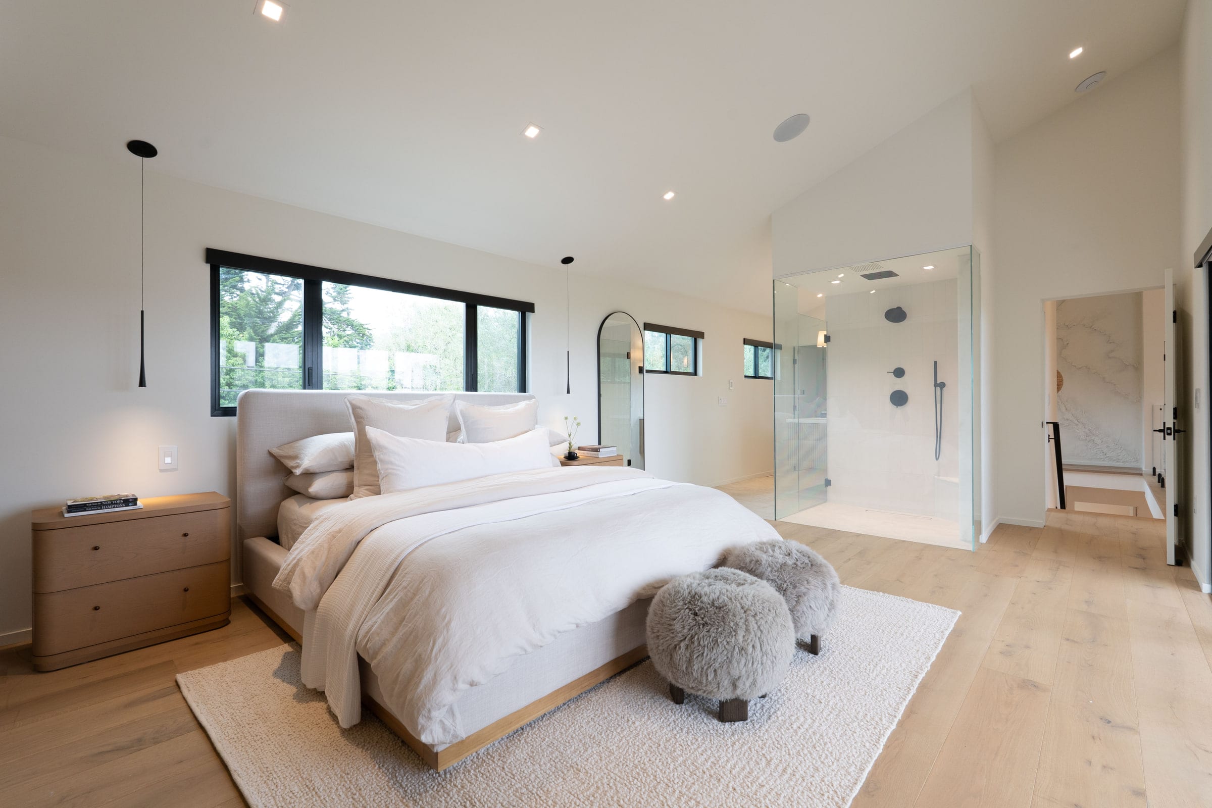 Primary bedroom with sheepskin stools at the foot of the bed and a glass-enclosed shower visible beyond, DHRH Padaro Lane renovation