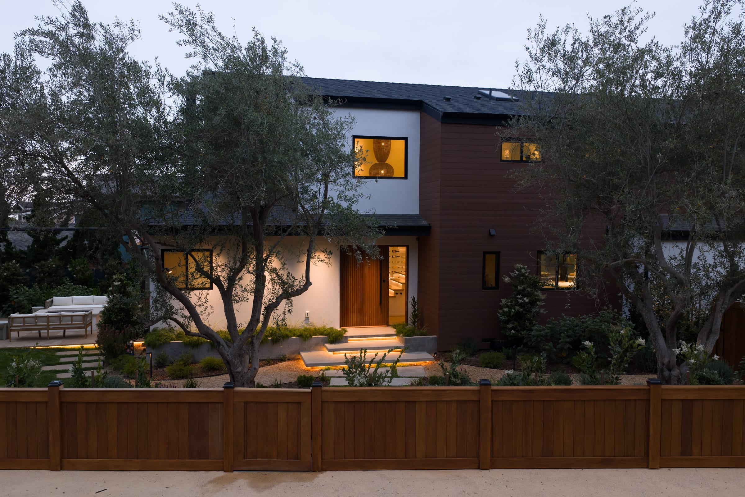 Twilight exterior of the two-story home with dark wood siding, white stucco, and a lit wood entry door, DHRH Padaro Lane renovation