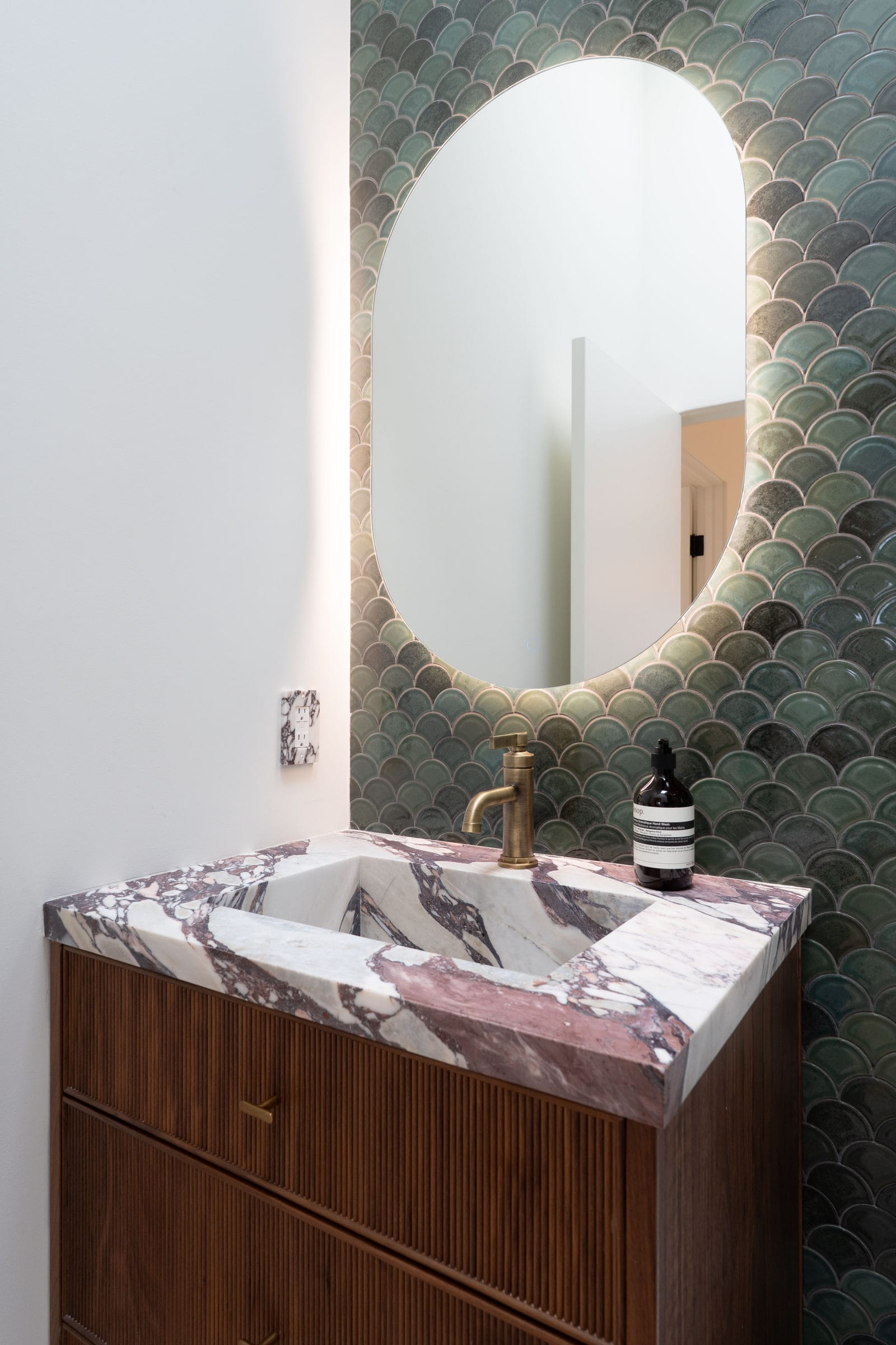 Close-up of a burgundy-veined marble vanity sink with a brass faucet and backlit oval mirror against green tile, DHRH Padaro Lane renovation