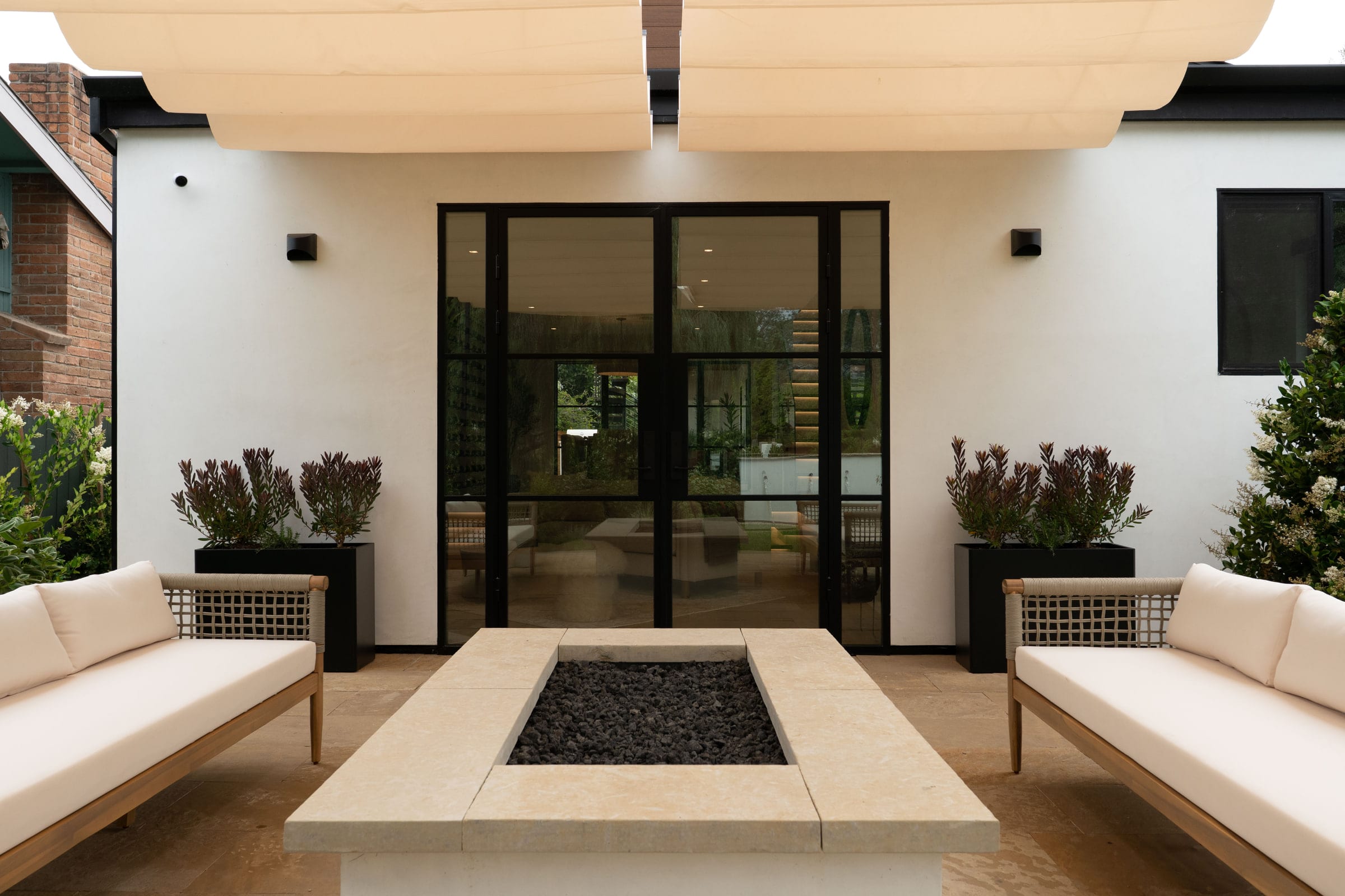 Outdoor lounge with cream sofas flanking a stone fire pit table under a canvas shade sail, DHRH Padaro Lane renovation