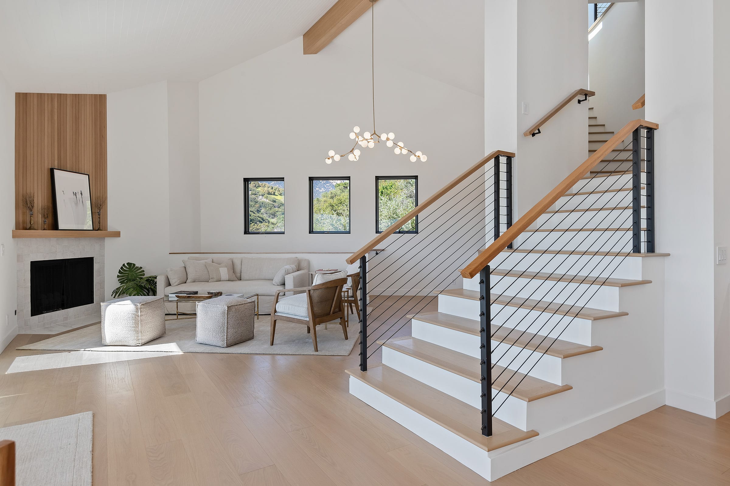 Open staircase with cable railings beside a living room with a branch chandelier and fireplace, DHRH Riviera renovation