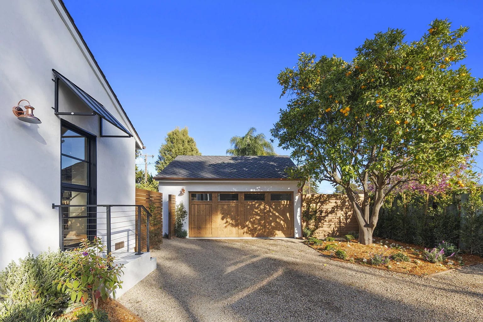 Exterior view of the wood carriage-style garage door and gravel driveway beside a citrus tree, DHRH San Roque renovation