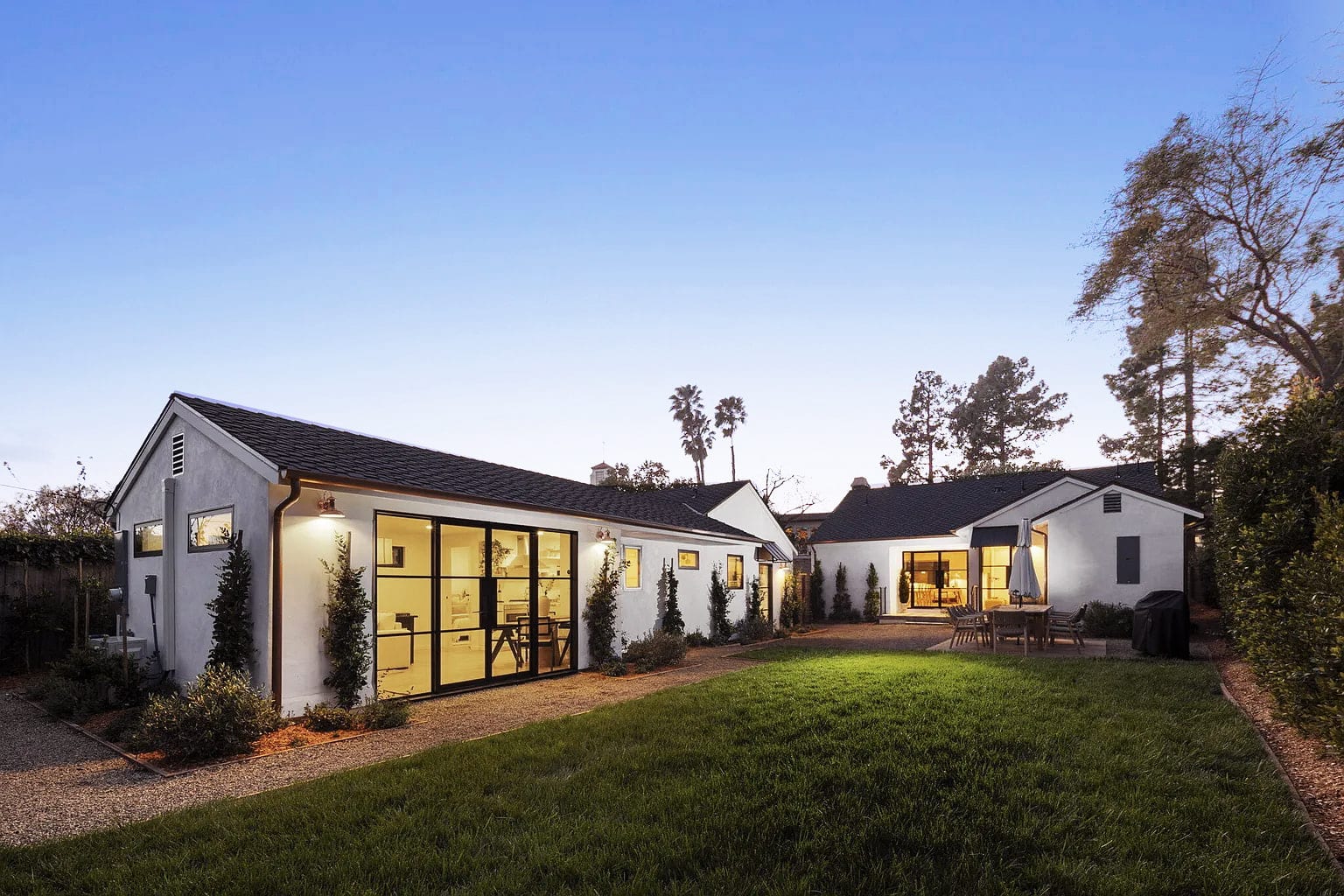 Twilight exterior of the single-story home with glowing black-framed glass doors and a gravel path, DHRH San Roque renovation