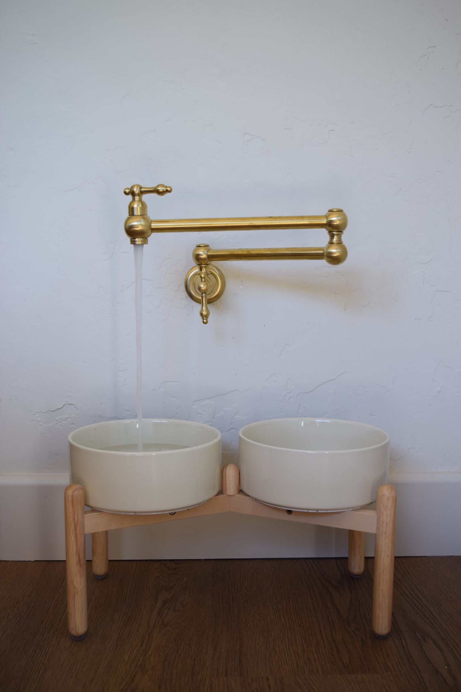 Close view of the wall-mounted brass faucet filling twin ceramic bowls on a raised wood dog-feeder stand, DHRH West Side renovation