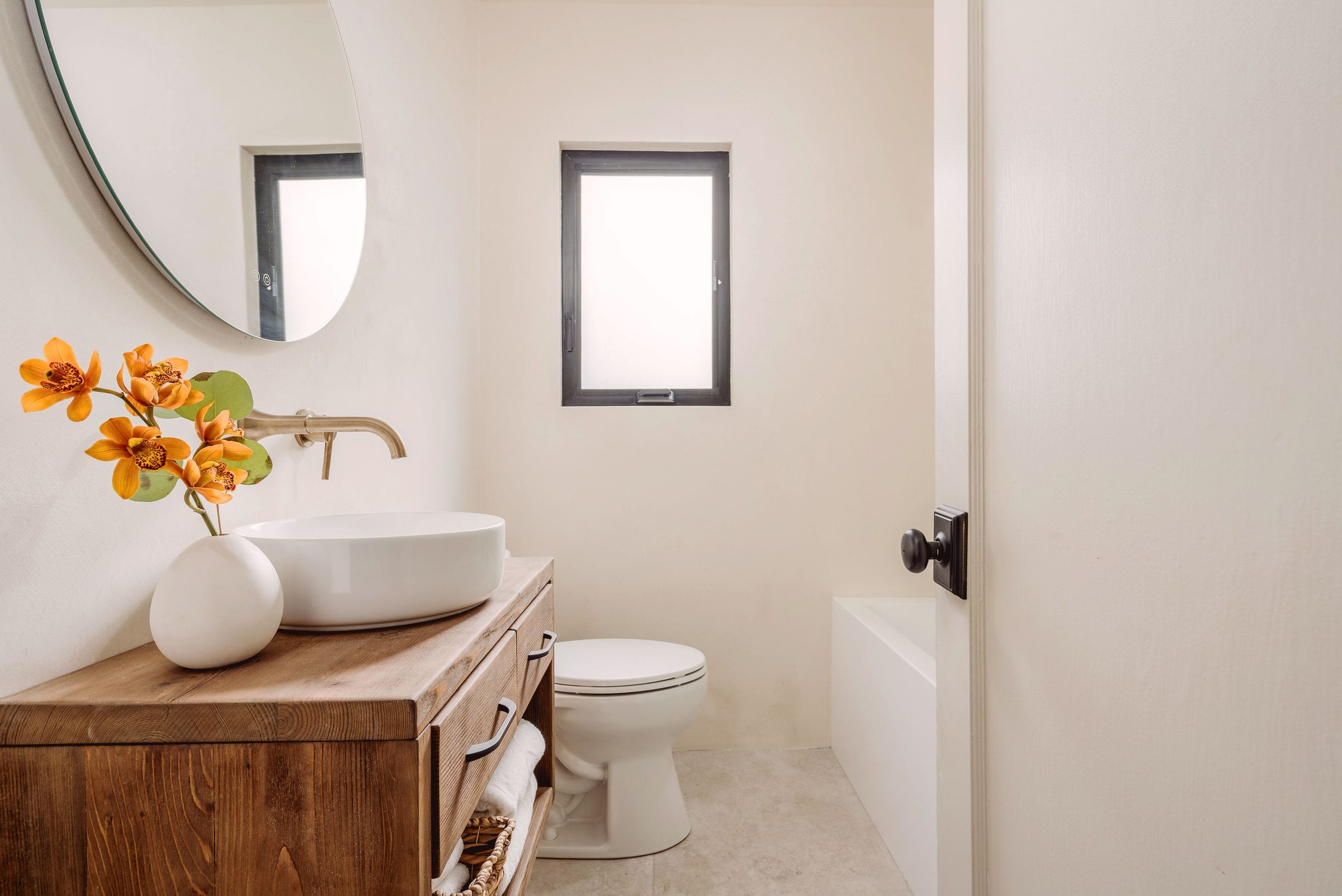 Powder room with a wood vanity, oval vessel sink, brass wall-mount faucet, and round arched mirror, DHRH West Side renovation