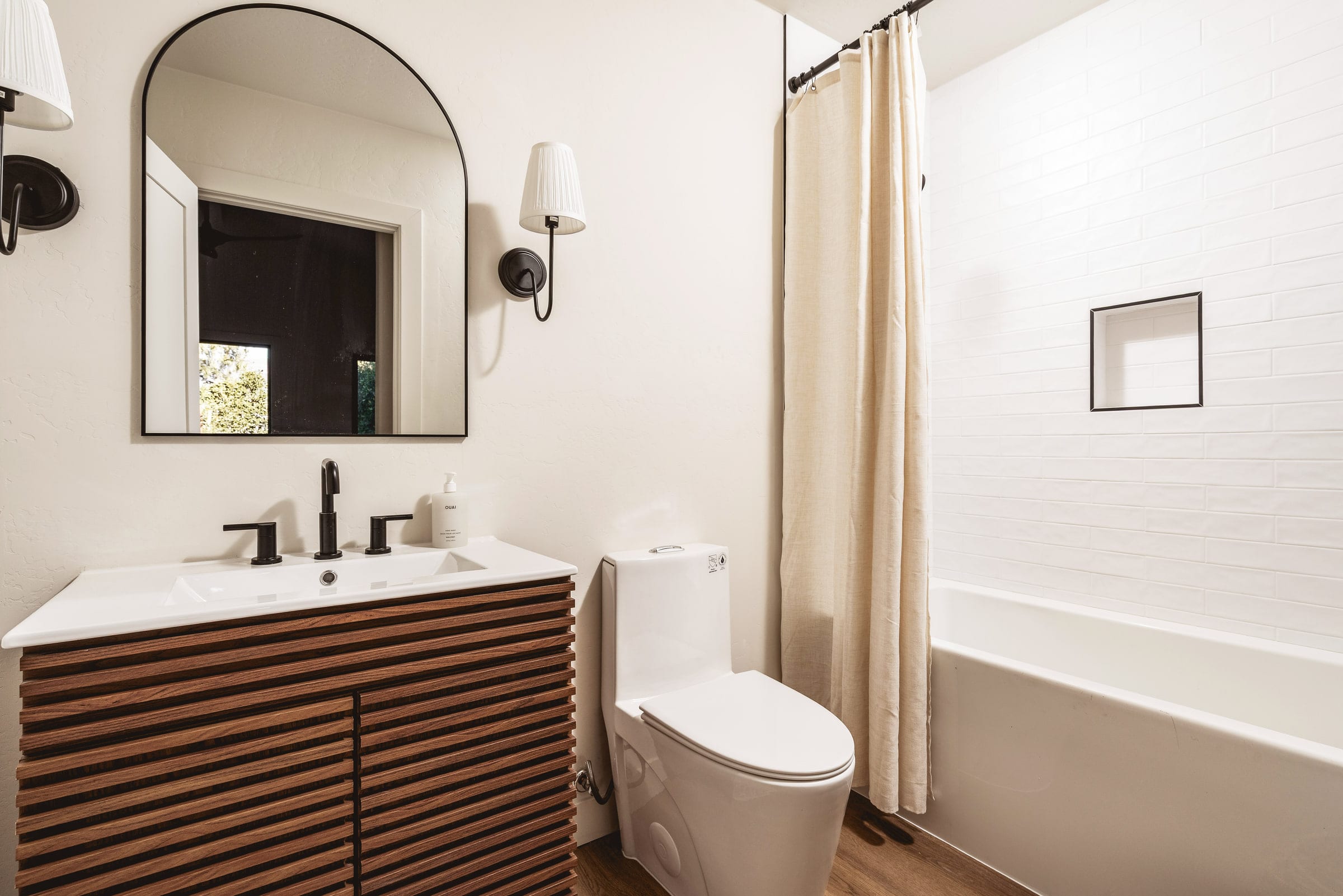 Bathroom with a wood slat vanity, arched mirror, black fixtures, and a white subway tile tub surround, DHRH West Side renovation
