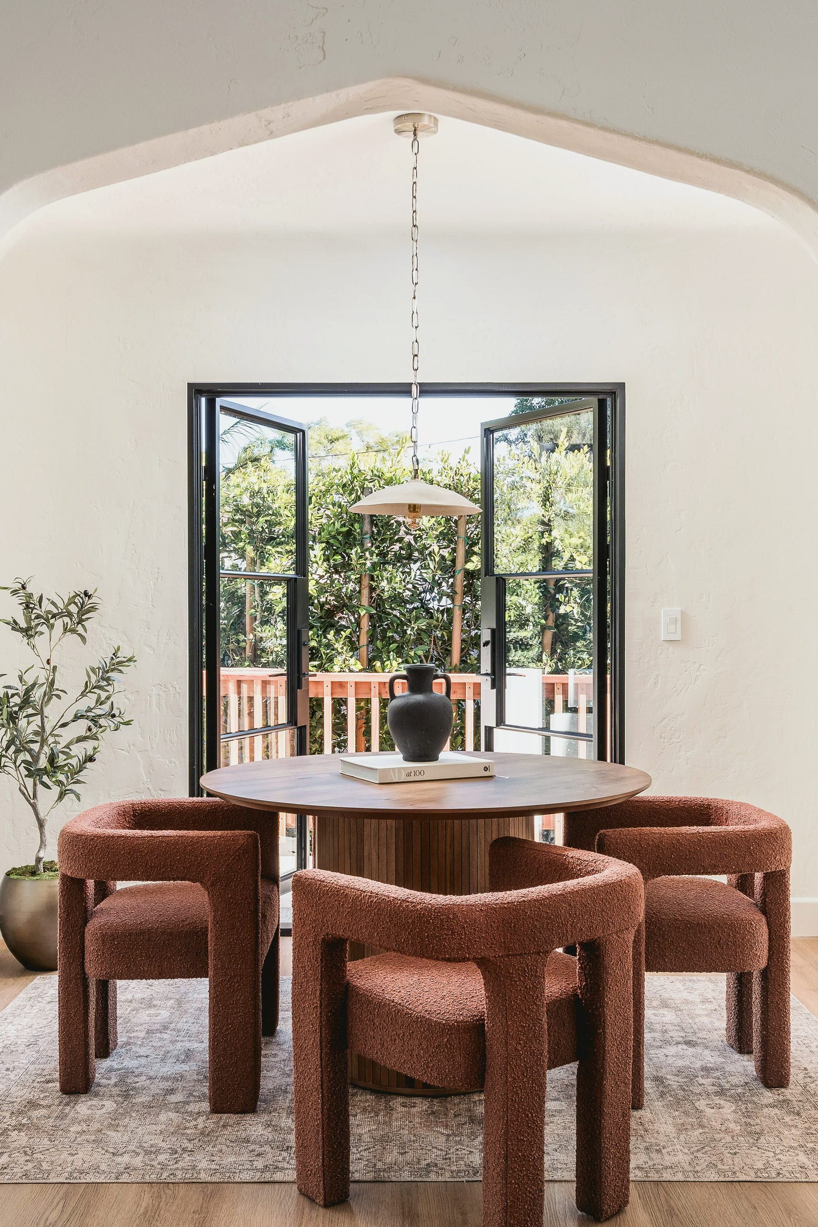 Round wood dining table with rust boucle chairs beneath a pendant light, opening to steel-framed garden doors, DHRH West Side renovation